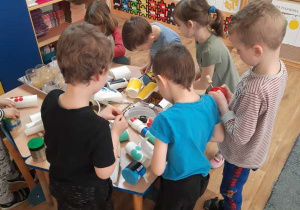 Dzieci stoją w sali wokół stołu i ozdabiają bębenki kolorowymi papierami.